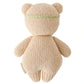 The Baby Honey Bear Ivory Floral by Cuddle + Kind is a collectible, heirloom-quality knitted teddy bear in beige, shown from the back and wearing a thin green headband.