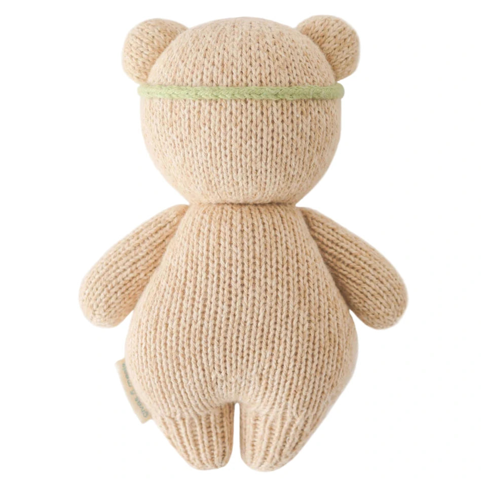 The Baby Honey Bear Ivory Floral by Cuddle + Kind is a collectible, heirloom-quality knitted teddy bear in beige, shown from the back and wearing a thin green headband.