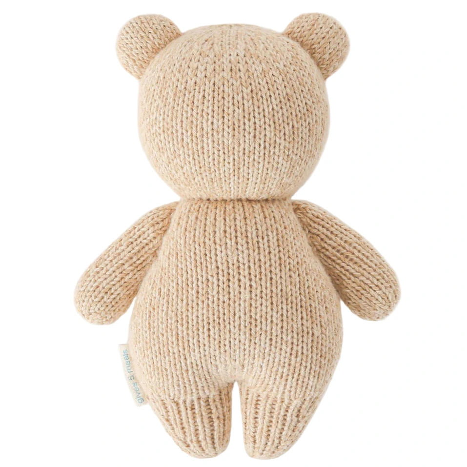 The Baby Honey Bear by Cuddle + Kind is a beige, hand-knit teddy bear made from heirloom-quality Peruvian cotton yarn, shown from the back with a round head, ears, arms, and legs but no visible facial features.