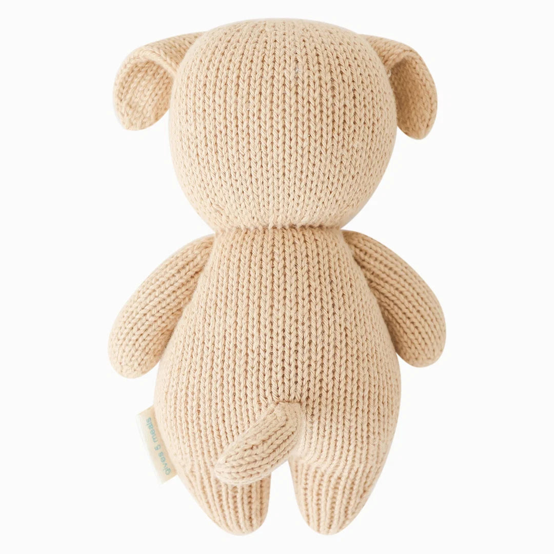 The Cuddle + Kind “Baby Puppy” is a handcrafted beige cotton plush toy shaped like a dog, shown from the back with floppy ears, a short tail, and a tag on its side. Designed to be a soft baby companion, it’s perfect for cuddles and gentle play.
