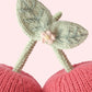 Close-up of the Baby Cherries by Cuddle + Kind—two meticulously handcrafted pink cherries with green stems and leaves, made from soft Peruvian cotton. A small pink knitted flower accents the leaf intersection, set against a light pink background. This heirloom-quality keepsake is the perfect soft companion for babies and little ones.