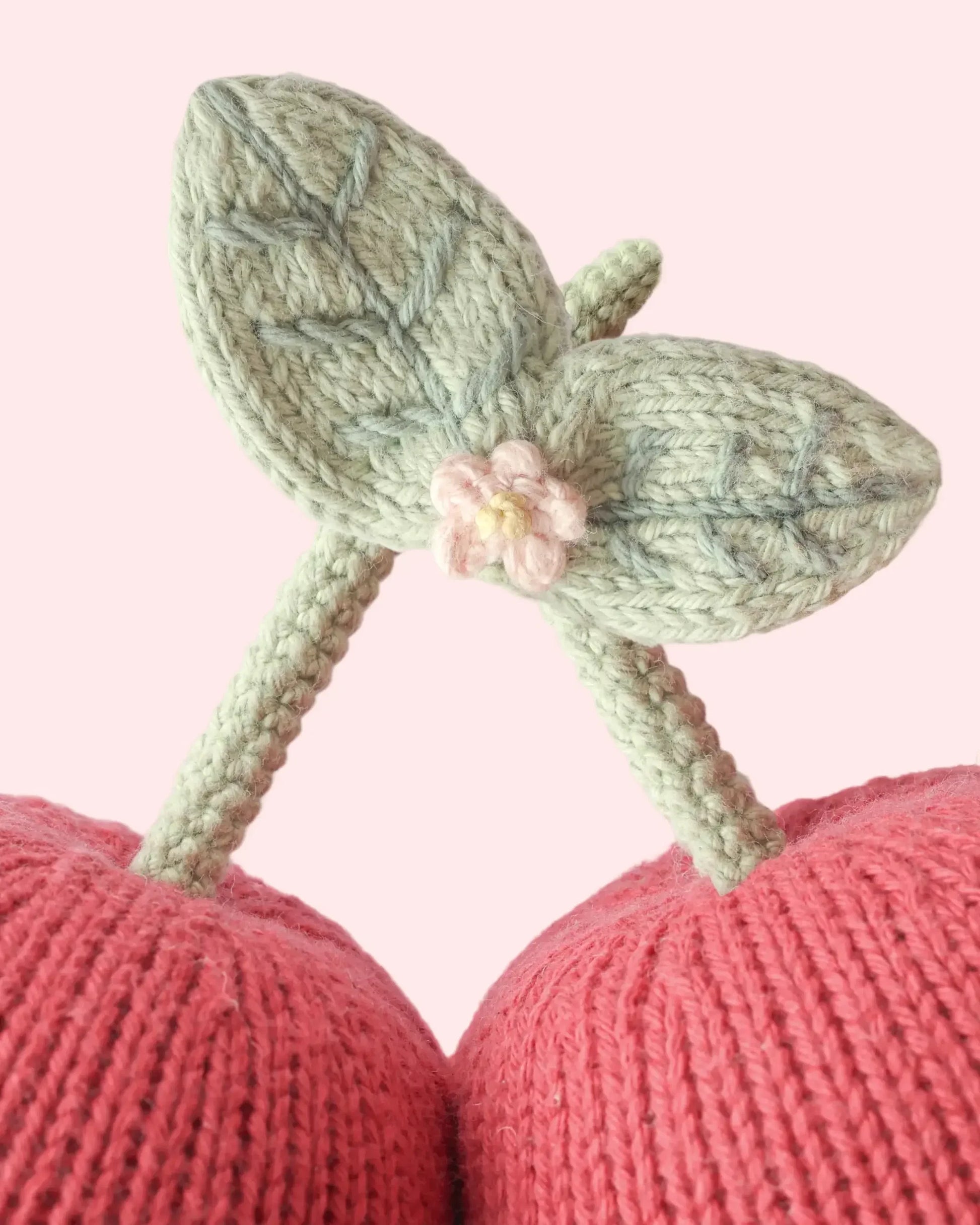 Close-up of the Baby Cherries by Cuddle + Kind—two meticulously handcrafted pink cherries with green stems and leaves, made from soft Peruvian cotton. A small pink knitted flower accents the leaf intersection, set against a light pink background. This heirloom-quality keepsake is the perfect soft companion for babies and little ones.