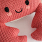Close-up of the Baby Crab Coral by Cuddle + Kind, a pink knitted crab plush toy crafted from handcrafted cotton yarn. This heirloom baby animal features black stitched eyes, a smiling mouth, and visible claws, making it a soft and cozy companion for little ones against a white background.