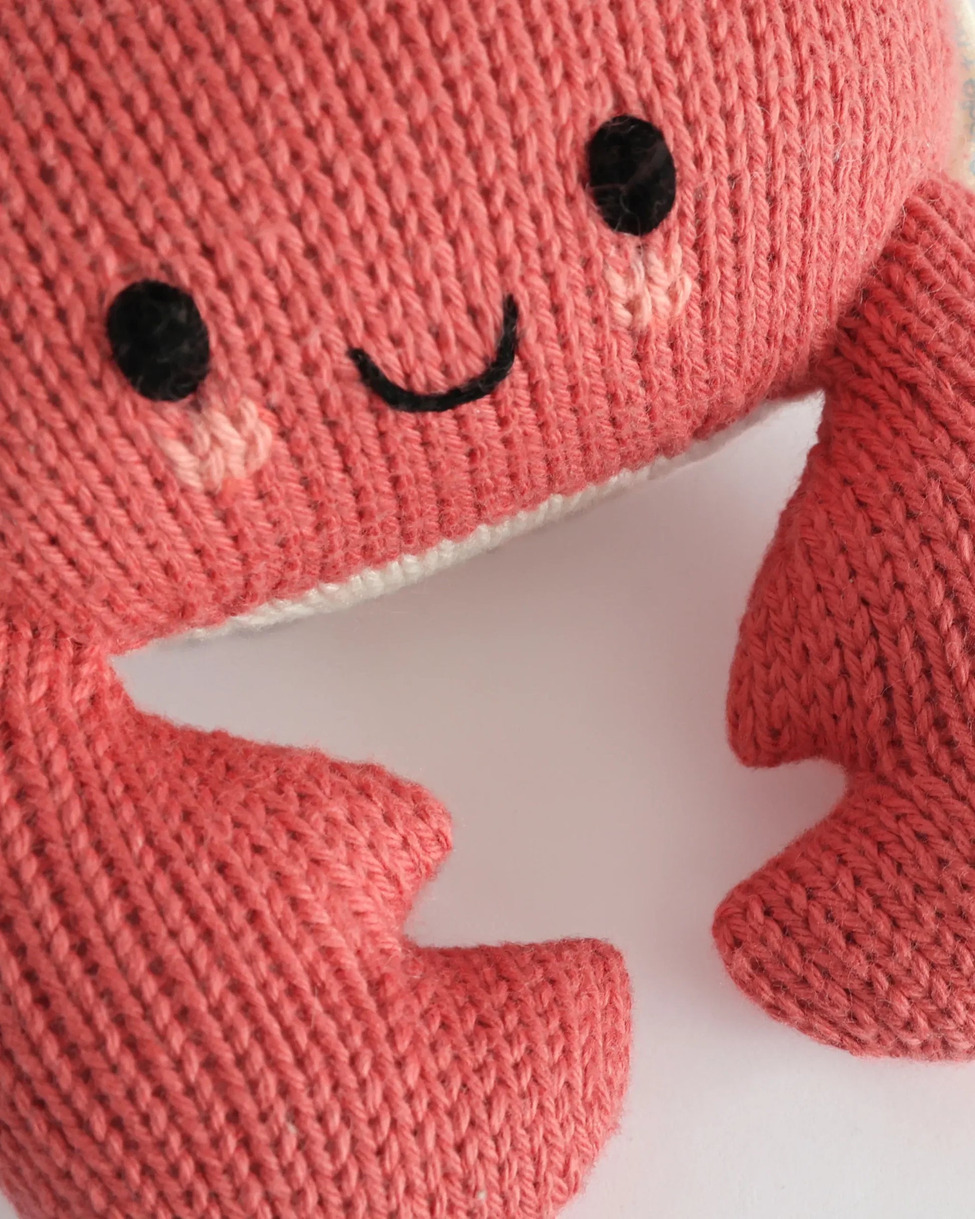 Close-up of the Baby Crab Coral by Cuddle + Kind, a pink knitted crab plush toy crafted from handcrafted cotton yarn. This heirloom baby animal features black stitched eyes, a smiling mouth, and visible claws, making it a soft and cozy companion for little ones against a white background.