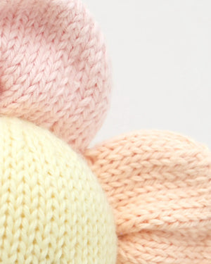 Close-up of three knitted fabrics in pastel colors—pink, yellow, and peach—arranged with the pink and peach pieces partially overlapping the yellow one. Each piece is crafted from soft, handcrafted cotton yarn by Cuddle + Kind, adding a gentle touch perfect for creating an heirloom Baby Daisy - Sunshower or a soft baby toy.