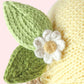 Close-up of the heirloom-quality Baby Lemon by Cuddle + Kind, expertly knitted from handcrafted Peruvian cotton in vibrant yellow, featuring two green leaves and a white flower with a yellow center, all made from yarn, against a pale pink background.