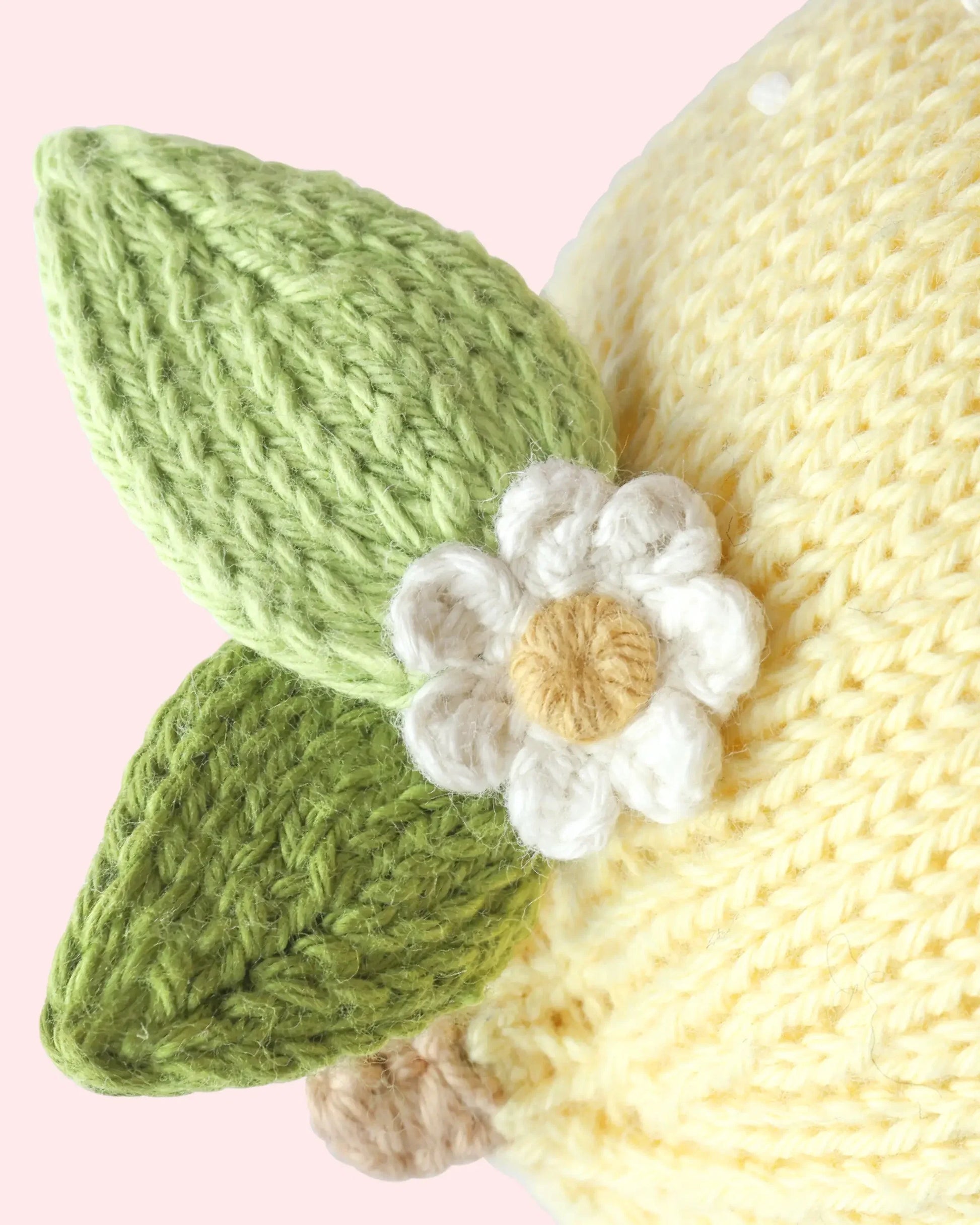 Close-up of the heirloom-quality Baby Lemon by Cuddle + Kind, expertly knitted from handcrafted Peruvian cotton in vibrant yellow, featuring two green leaves and a white flower with a yellow center, all made from yarn, against a pale pink background.