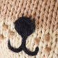 Close-up of beige knitted fabric handcrafted from Peruvian cotton, featuring a black, upside-down “Y” shaped stitched embroidery on the Baby Otter by Cuddle + Kind.