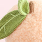 Close-up of the Baby Peach plush toy by Cuddle + Kind, handcrafted from Peruvian cotton yarn with a textured surface. It features two green knitted leaves and a short brown stem, set against a light pink background.