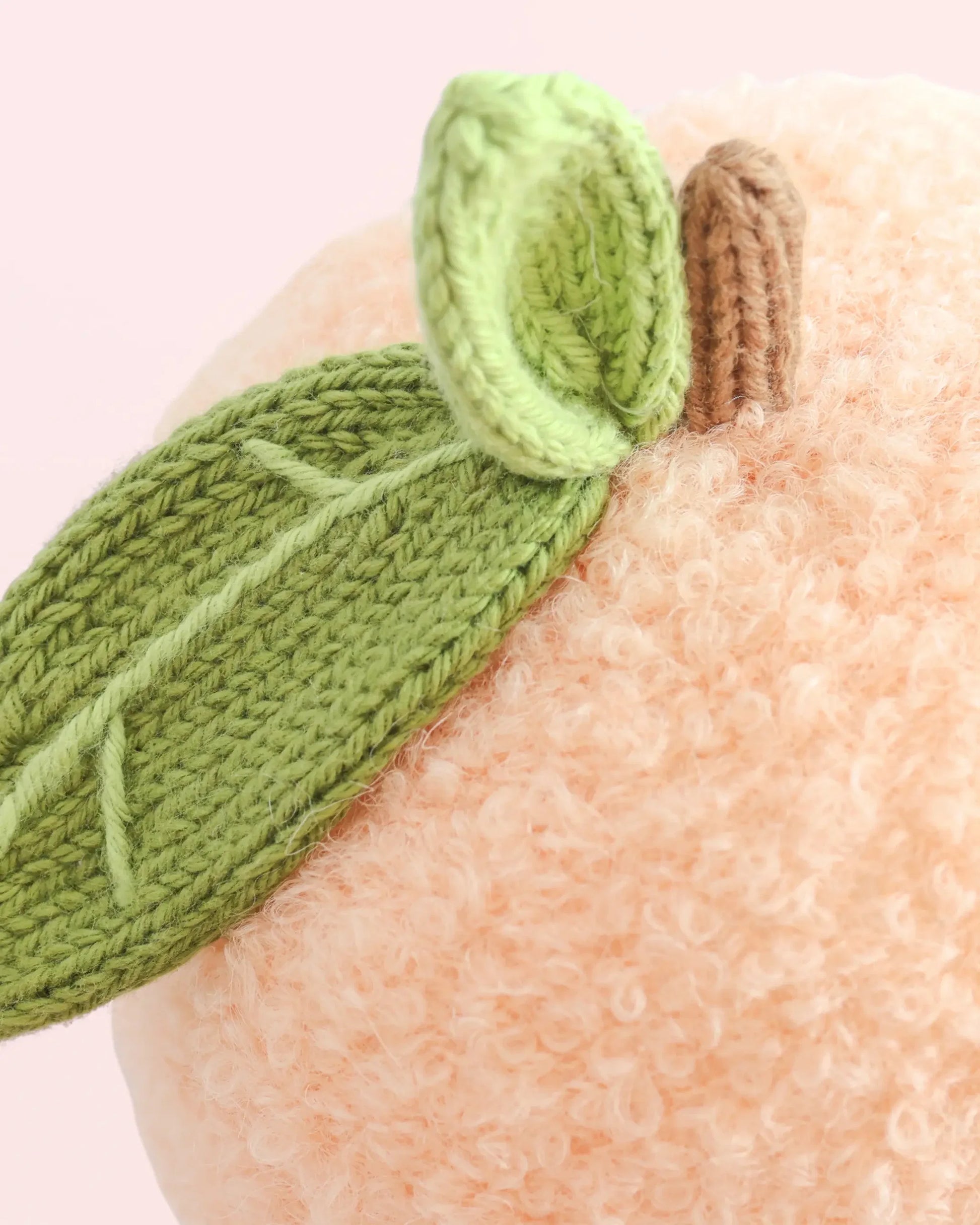 Close-up of the Baby Peach plush toy by Cuddle + Kind, handcrafted from Peruvian cotton yarn with a textured surface. It features two green knitted leaves and a short brown stem, set against a light pink background.