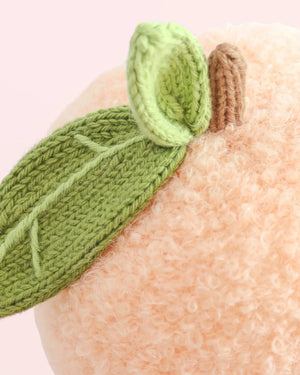 Close-up of the Baby Peach plush toy by Cuddle + Kind, handcrafted from Peruvian cotton yarn with a textured surface. It features two green knitted leaves and a short brown stem, set against a light pink background.