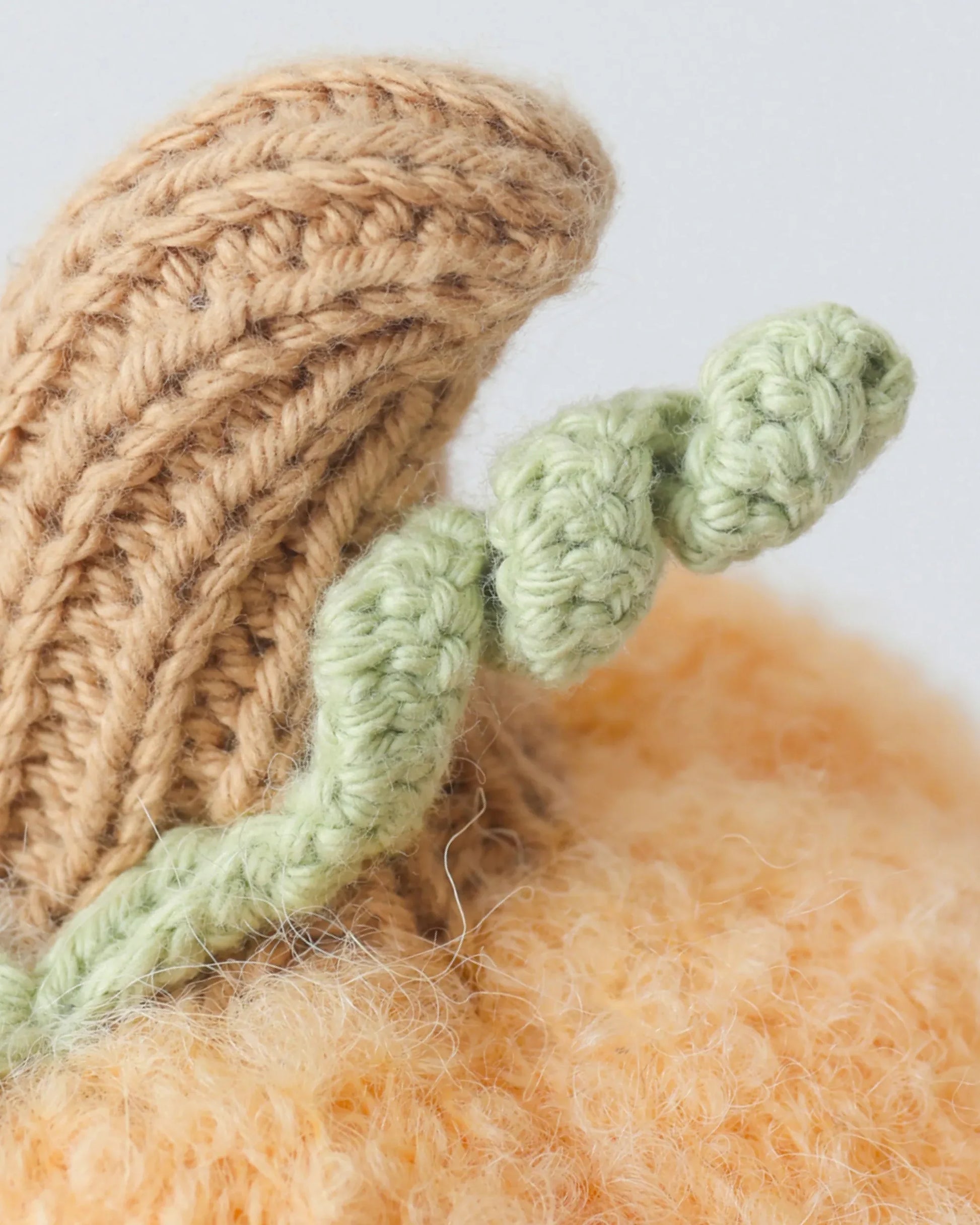 Close-up of the Baby Pumpkin by Cuddle + Kind, a knitted object featuring a brown stem and green, twisted vine detail, lovingly handcrafted from soft cotton yarn—an ideal companion for your baby.