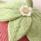 Close-up of the Cuddle + Kind Baby Strawberry, a handcrafted item knitted from soft Peruvian cotton yarn, featuring green leaves and a white flower reminiscent of a strawberry top, set against a textured pink background.
