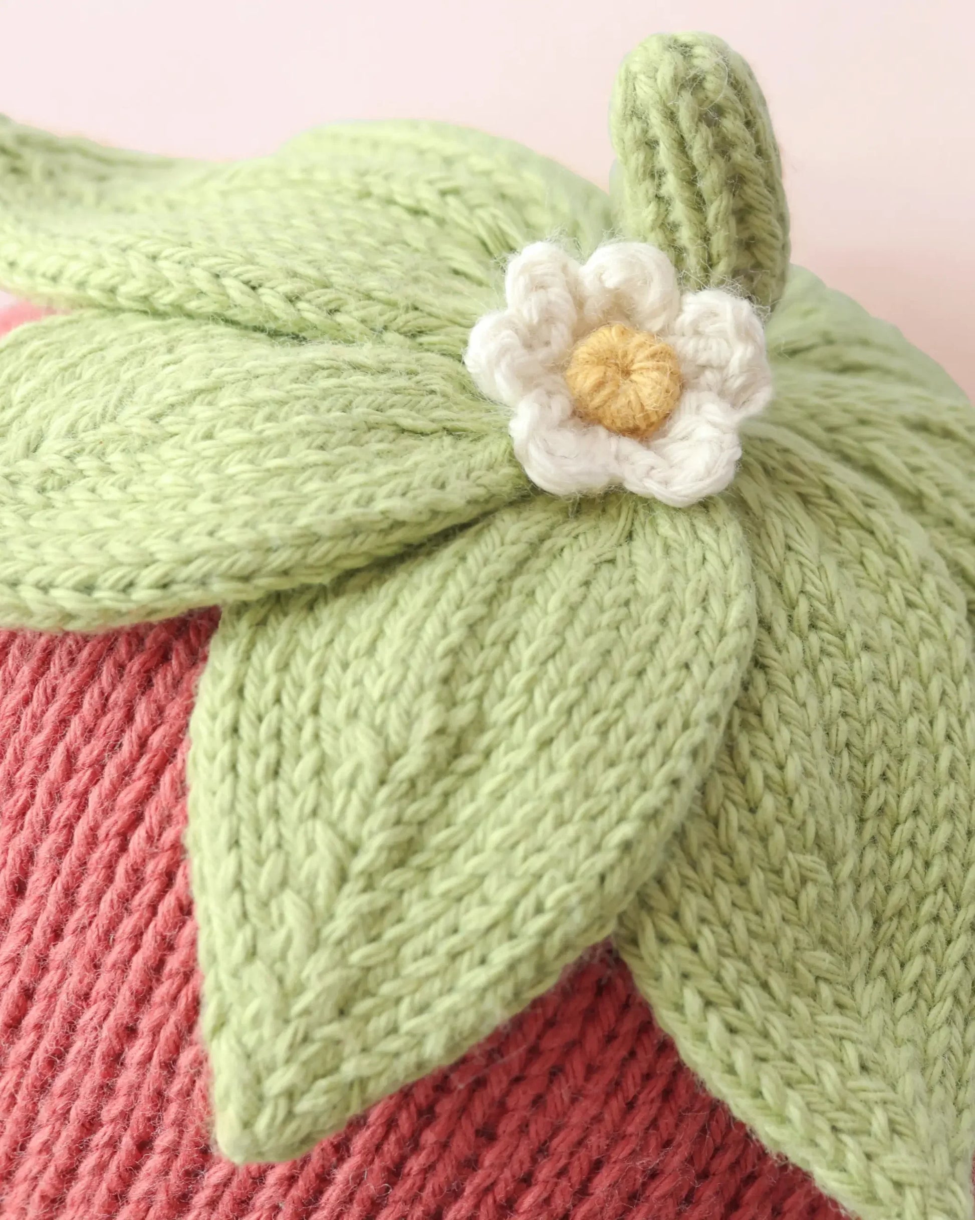 Close-up of the Cuddle + Kind Baby Strawberry, a handcrafted item knitted from soft Peruvian cotton yarn, featuring green leaves and a white flower reminiscent of a strawberry top, set against a textured pink background.