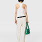 A person with short dark hair, shown from the back against a plain background, wears a white tank top, Stella McCartney Washed Cotton Straight Trousers, black shoes and belt, and holds a green handbag.
