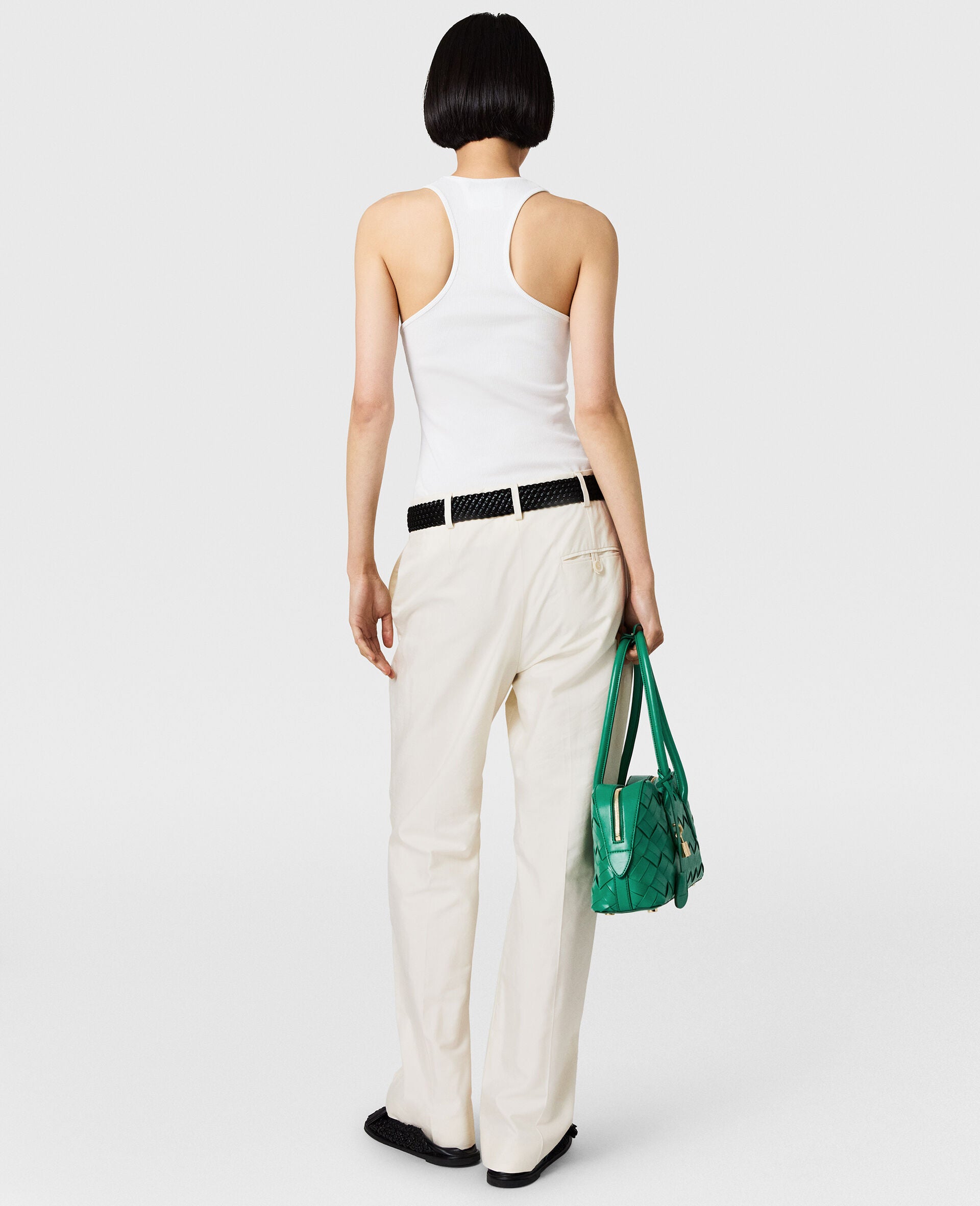 A person with short dark hair, shown from the back against a plain background, wears a white tank top, Stella McCartney Washed Cotton Straight Trousers, black shoes and belt, and holds a green handbag.