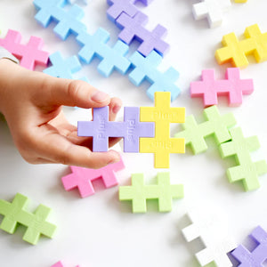 A hand holds two interlocking Plus Plus Tube - BIG 15 Piece - Pastel blocks above a scattered assortment, encouraging creative play and enhancing fine motor skills.