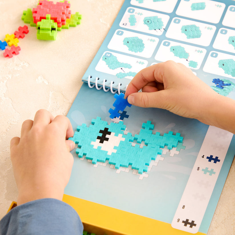A child assembles a blue pixel-style fish puzzle on a table, following illustrated instructions from the spiral-bound guidebook included in the Plus Plus Puzzle By Number® Activity Pad—an engaging experience that fosters creativity and enhances fine motor skills.
