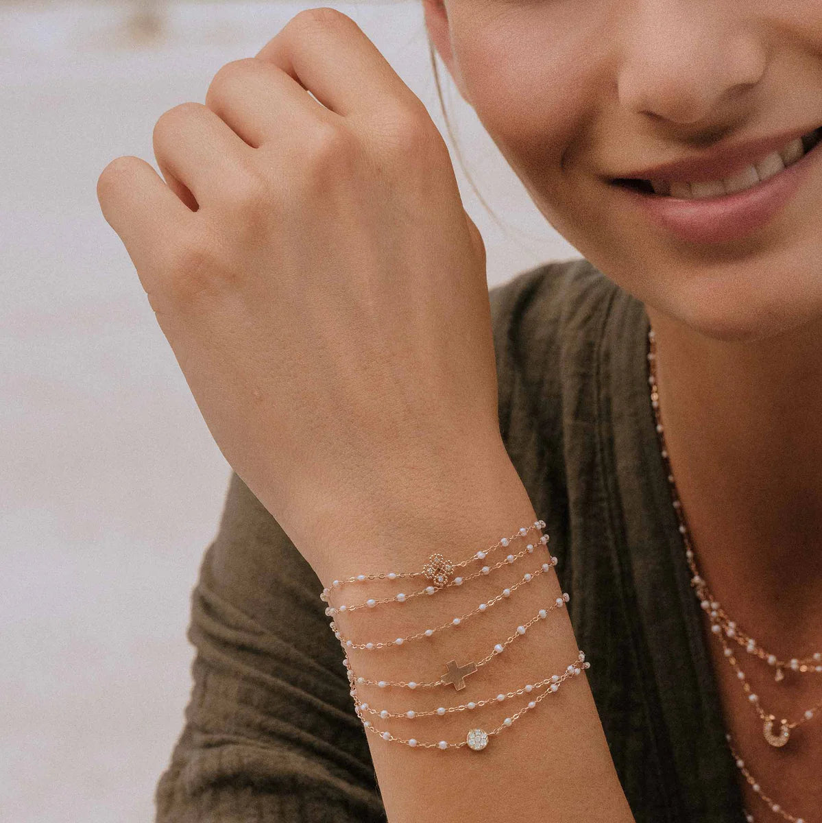 A person wears multiple delicate 18k gold and pearl bracelets, including the Gigi Clozeau Classic Cross Charm Bracelet 6.7" with elegant resin pearls, their wrist and a soft smile partly visible.