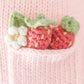 Ava the Cow Powder Pink - Little 13" by Cuddle + Kind features two knitted strawberries and a white flower tucked inside a pink knitted pocket, making it a sweet symbol of friendship or an adorable birthday gift.