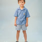 A young boy stands against a light background, smiling slightly with his hands by his sides, wearing a blue short-sleeve button-up shirt, Gingersnaps Boys' Pablo Twill Baltik Tile Drawstring Shorts, and black sandals.