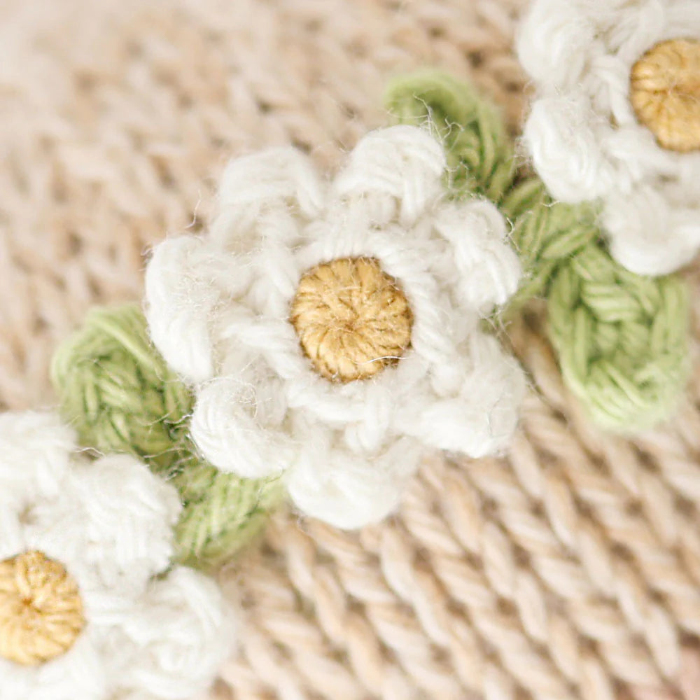 Close-up of Cuddle + Kind’s Baby Honey Bear Ivory Floral, featuring heirloom-quality crocheted white flowers with yellow centers and green leaves on a beige knitted background.