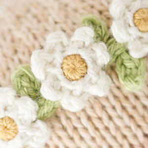 Close-up of Cuddle + Kind’s Baby Honey Bear Ivory Floral, featuring heirloom-quality crocheted white flowers with yellow centers and green leaves on a beige knitted background.