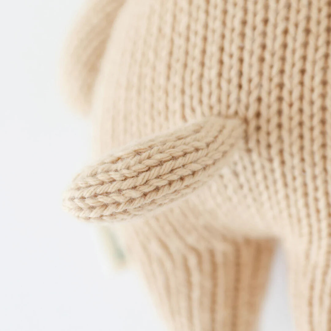 Close-up of the Cuddle + Kind “Baby Puppy,” a knitted beige stuffed animal handcrafted from cotton yarn, showing its tail and part of its legs, photographed against a plain white background.