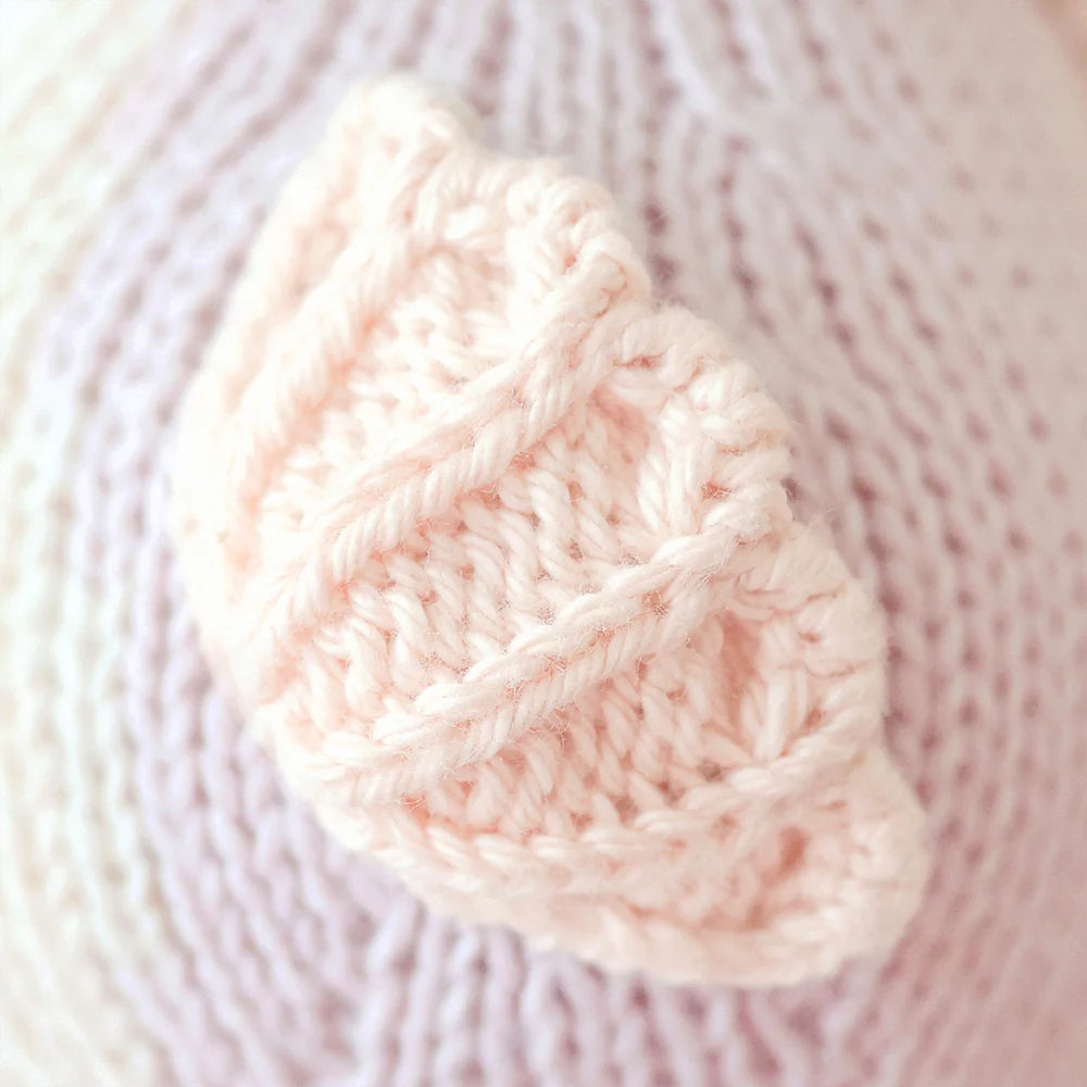 Close-up of the Cuddle + Kind Baby Seahorse Lilac, featuring a pale pink leaf knit pattern, exquisitely crafted with heirloom-quality Peruvian cotton yarn and resting on a matching knitted fabric background.
