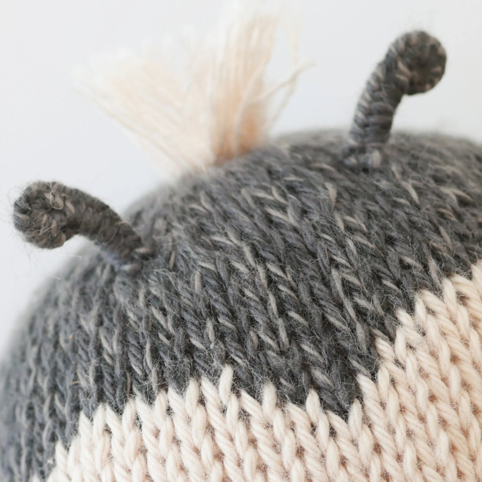 Close-up of the Cuddle + Kind Baby Ladybug, a collectible hand-knit baby animal made with gray and cream yarn, featuring two short, curled antennae and a tuft of cream yarn on top.