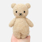 A hand holds the Cuddle + Kind Baby Boucle Bear - Honey, a plush beige teddy made from heirloom-quality Peruvian cotton yarn with black eyes, a black nose, and embroidered blush cheeks, shown against a plain white background.