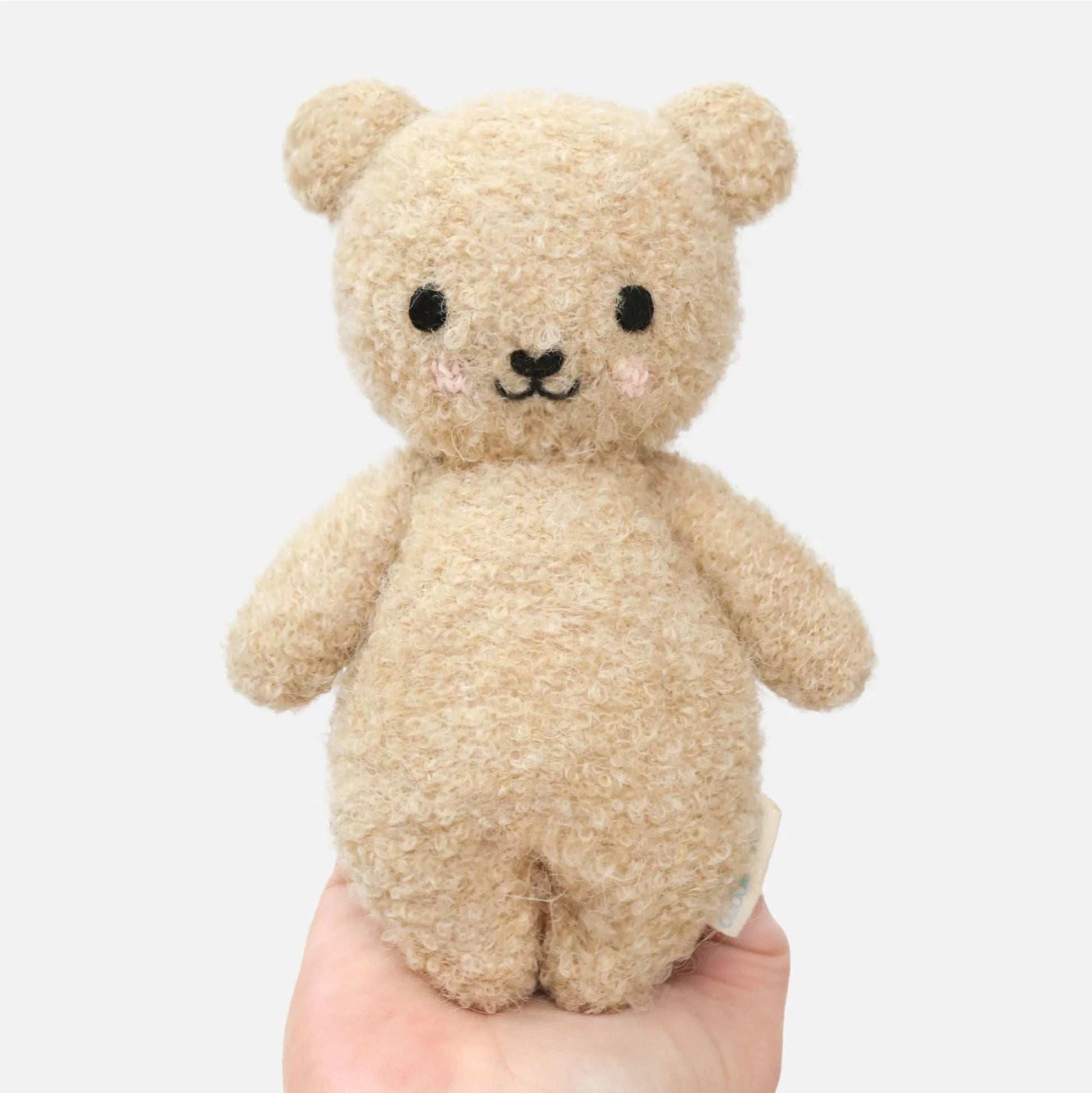 A hand holds the Cuddle + Kind Baby Boucle Bear - Honey, a plush beige teddy made from heirloom-quality Peruvian cotton yarn with black eyes, a black nose, and embroidered blush cheeks, shown against a plain white background.