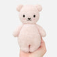 A hand holds the Cuddle + Kind Baby Boucle Bear - Strawberry, a small, light pink heirloom-quality plush teddy bear with black eyes, a black nose, and a simple smile, against a plain white background.