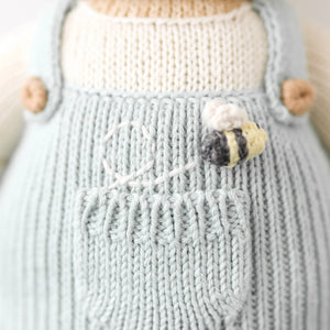 Close-up of Charlie the Honey Bear by Cuddle + Kind—a sweet knitted bear in light blue overalls with a bee and white flower embroidered on the front. Regular size, 20".