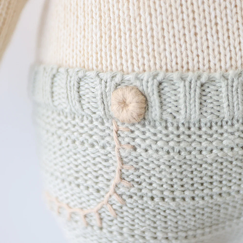 Close-up of Sawyer the Lion - Regular 20" by Cuddle + Kind shows cream and light gray knitted fabric, a round button, and beige stitching—a thoughtfully textured plush that's perfect as a birthday gift.
