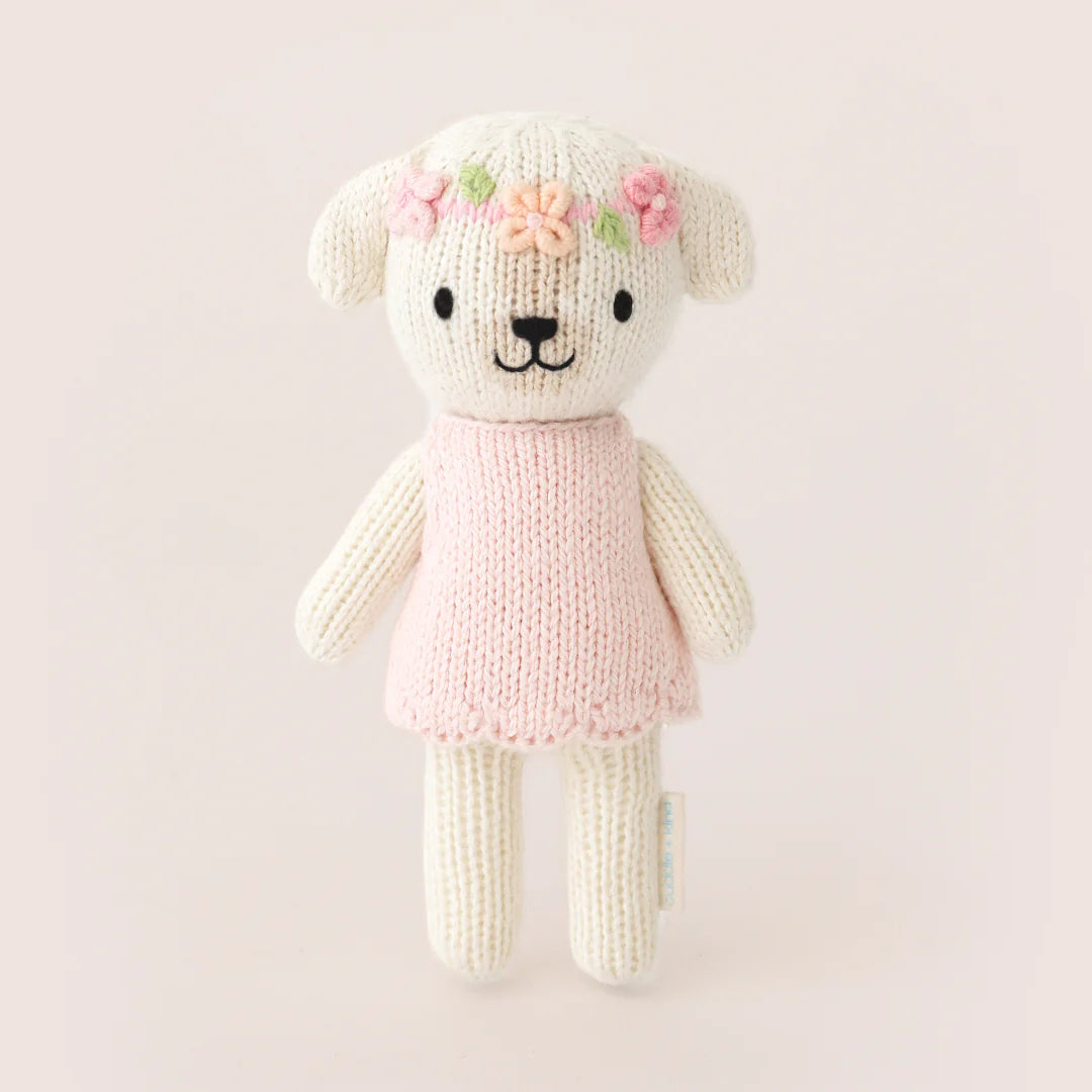 Tiny Charlotte the Dog by Cuddle + Kind is a hand-knit plush from the Tiny Collection, dressed in a pink dress and flower crown, shown standing upright against a simple light background. Perfect for collectors and snuggles.