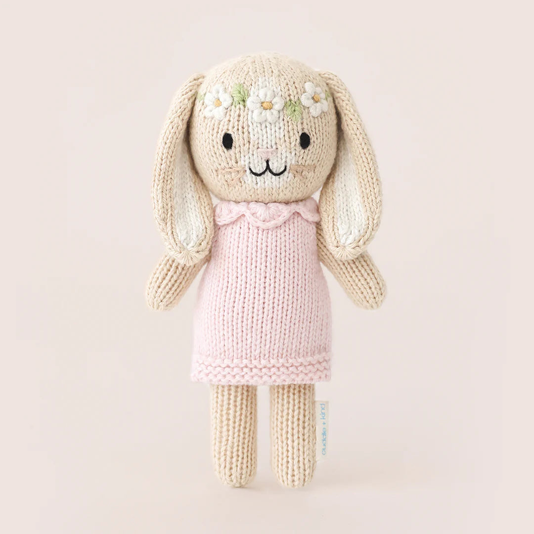Tiny Hannah the Bunny Blush by Cuddle + Kind is a fair trade, hand-knit doll with long ears, a pink dress, and a flower crown, standing against a plain light background.