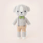 The Cuddle + Kind Tiny Noah the Dog, a hand-knit small dog doll in gray and white yarn with tan shorts, a green bow tie, and suspenders, stands upright against a plain light background.