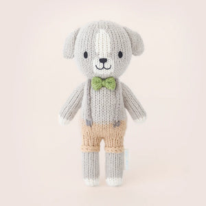 The Cuddle + Kind Tiny Noah the Dog, a hand-knit small dog doll in gray and white yarn with tan shorts, a green bow tie, and suspenders, stands upright against a plain light background.