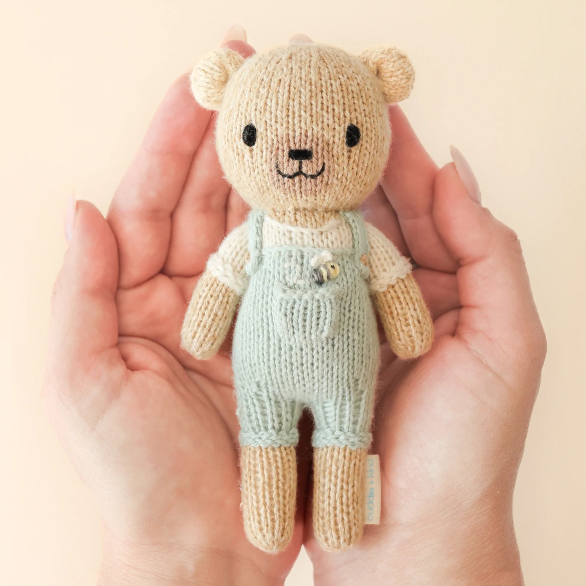 A pair of hands holds Tiny Charlie the Honey Bear from Cuddle + Kind—a hand-knit, fair trade teddy bear in pale green overalls and a cream shirt—against a light beige background.