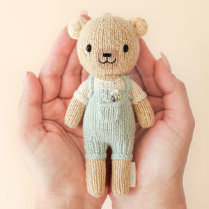 A pair of hands holds Tiny Charlie the Honey Bear from Cuddle + Kind—a hand-knit, fair trade teddy bear in pale green overalls and a cream shirt—against a light beige background.