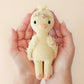 Two hands gently hold a Cuddle + Kind Tiny Flora the Duckling Mauve plush toy with a flower crown on its head, against a light beige background.