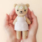 Two hands hold Cuddle + Kind’s Tiny Olivia the Honey Bear, a hand-knit doll in a white apron and flower crown, against a beige background.