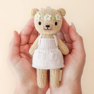 Two hands hold Cuddle + Kind’s Tiny Olivia the Honey Bear, a hand-knit doll in a white apron and flower crown, against a beige background.