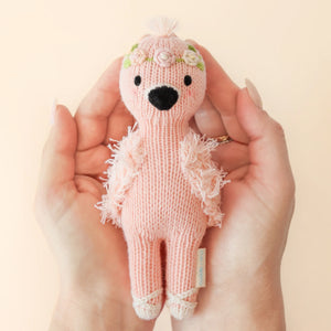 Two hands hold Tiny Penelope the Flamingo—a hand-knit pink doll with a black beak, floral headband, and textured wings—from Cuddle + Kind. The soft toy is shown against a pale background.