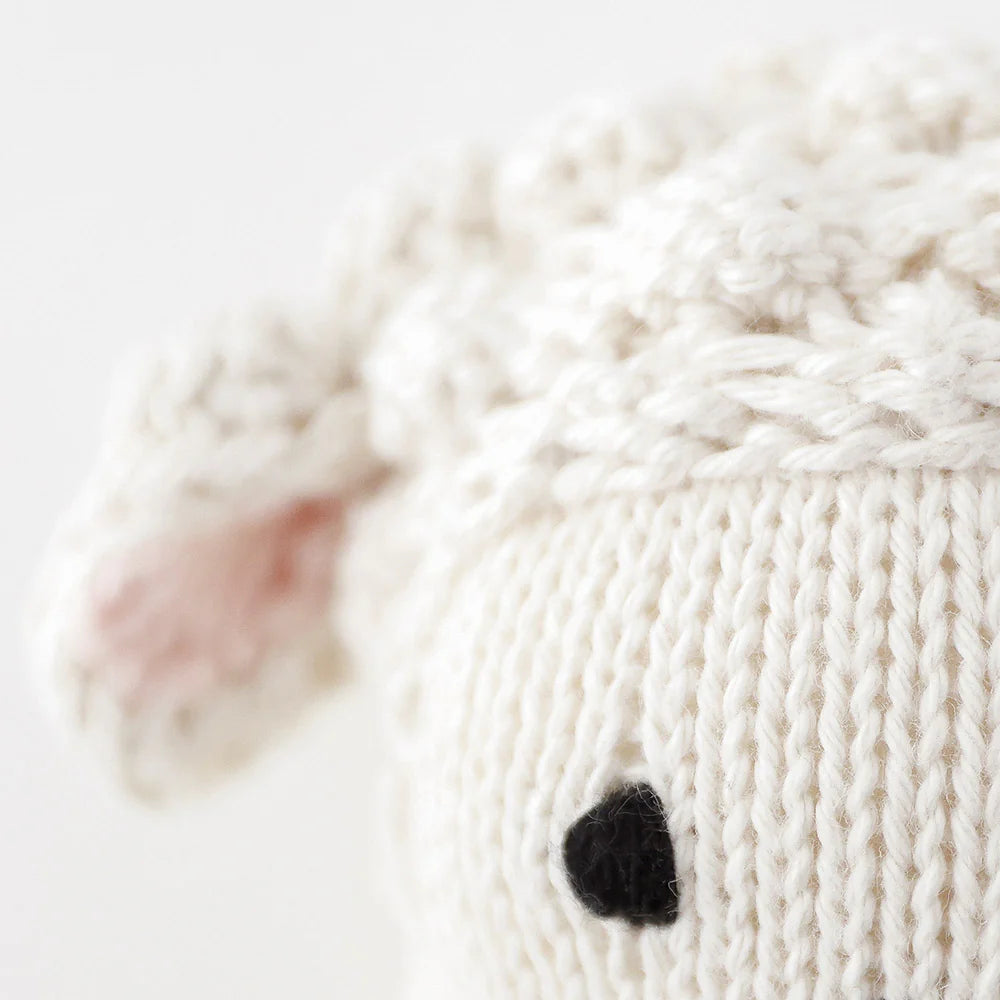 Close-up of the Baby Lamb by Cuddle + Kind, featuring handcrafted cotton yarn, a soft white head, pink ear detail, and black stitched eye—an adorable plush for any collector.