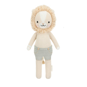 Sawyer the Lion - Little 13" by Cuddle + Kind is a knitted plush toy featuring a beige body, fluffy mane, embroidered face, and light gray shorts—beautifully crafted to inspire courage and kindness in every hug, shown against a white background.