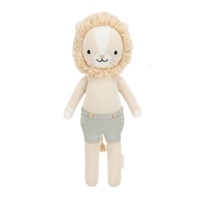 Sawyer the Lion - Little 13" by Cuddle + Kind is a knitted plush toy featuring a beige body, fluffy mane, embroidered face, and light gray shorts—beautifully crafted to inspire courage and kindness in every hug, shown against a white background.
