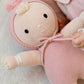 A Cuddle + Kind My First Baby doll, hand-knit from natural cotton, lies on a soft surface beside a baby’s arm. The doll features a light hair tuft, a pink hood with pom-pom, and a hospital wristband on its arm.