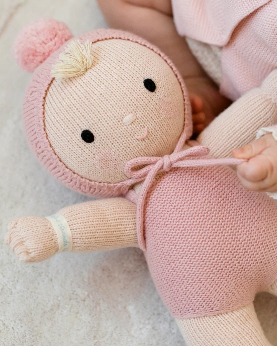 A Cuddle + Kind My First Baby doll, hand-knit from natural cotton, lies on a soft surface beside a baby’s arm. The doll features a light hair tuft, a pink hood with pom-pom, and a hospital wristband on its arm.